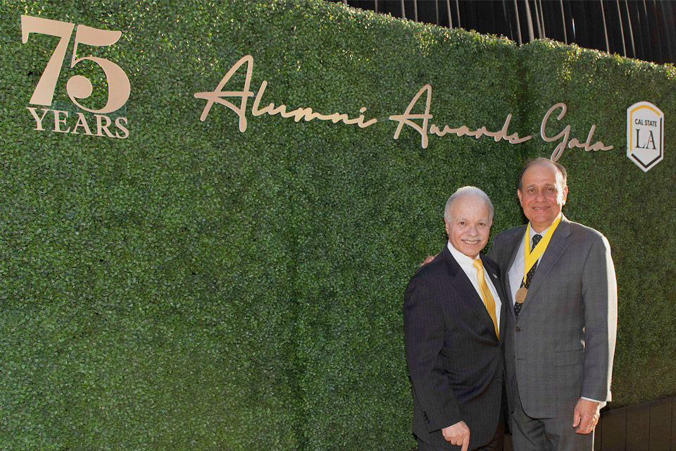 President Covino and alumnus Willie Zuniga in front of a decorative hedge adorned with gold writing that says 75 years, Alumni Awards Gala.
