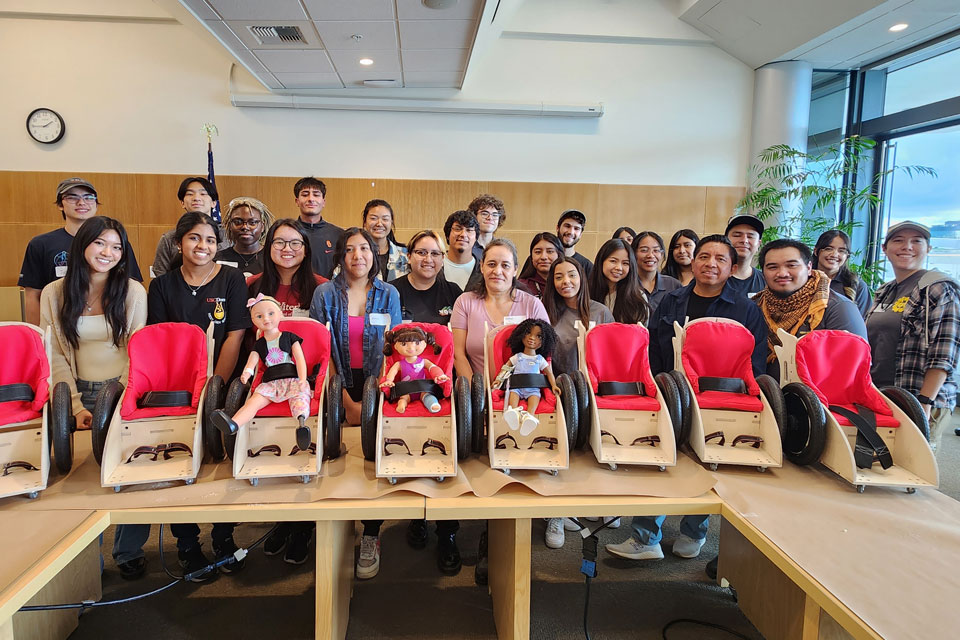 A large group of students posing for a photo in front toy dolls placed in child-sized seats.