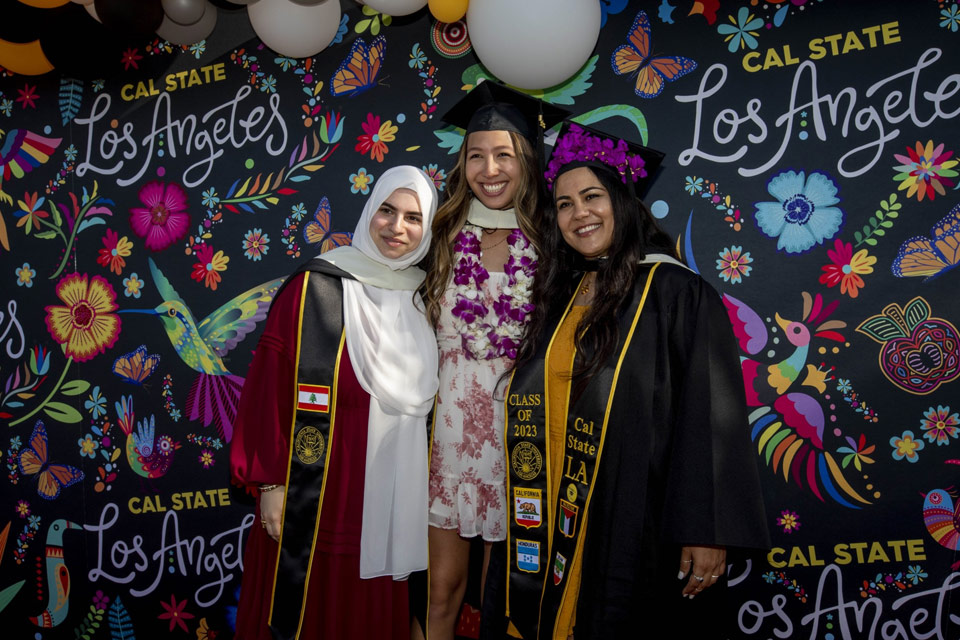 Three Cal State LA graduates taking a photo together in front of a colorful backdrop.