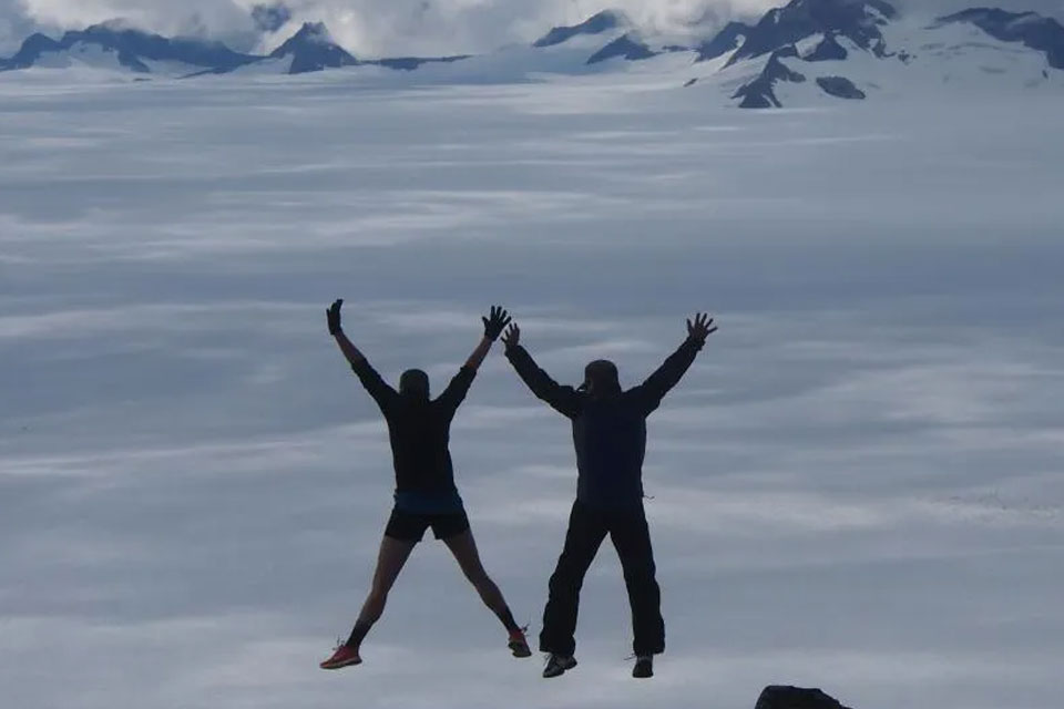 Two people jumping high and stretching out their arms and legs. Snow mountains are in the background.