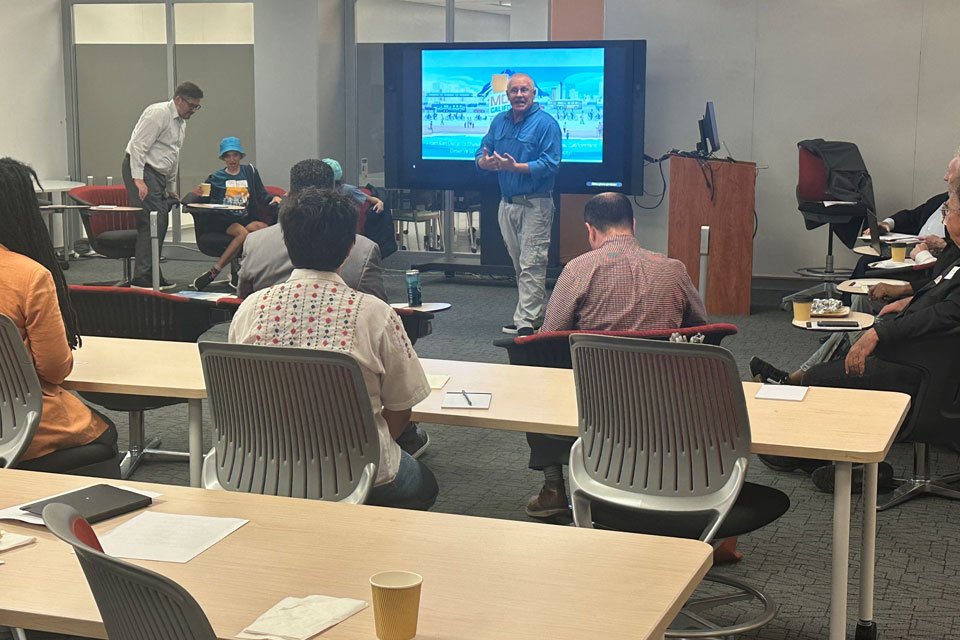 Indoor presentation with Move LA attendees seated in a classroom setting, listening to a speaker in front of a screen displaying a slide deck.