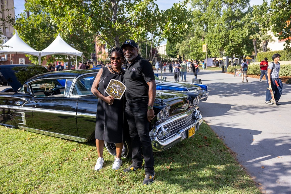 Two people smile in front of a black classic Chevrolet parked on the Cal State LA campus during Campus Day.