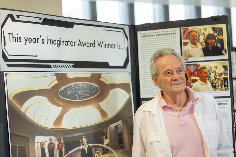 Imaginator Award winner Herman Zimmerman stands beside a large poster exhibit honoring his career in production design, on display at Cal State LA’s Eagle-Con.