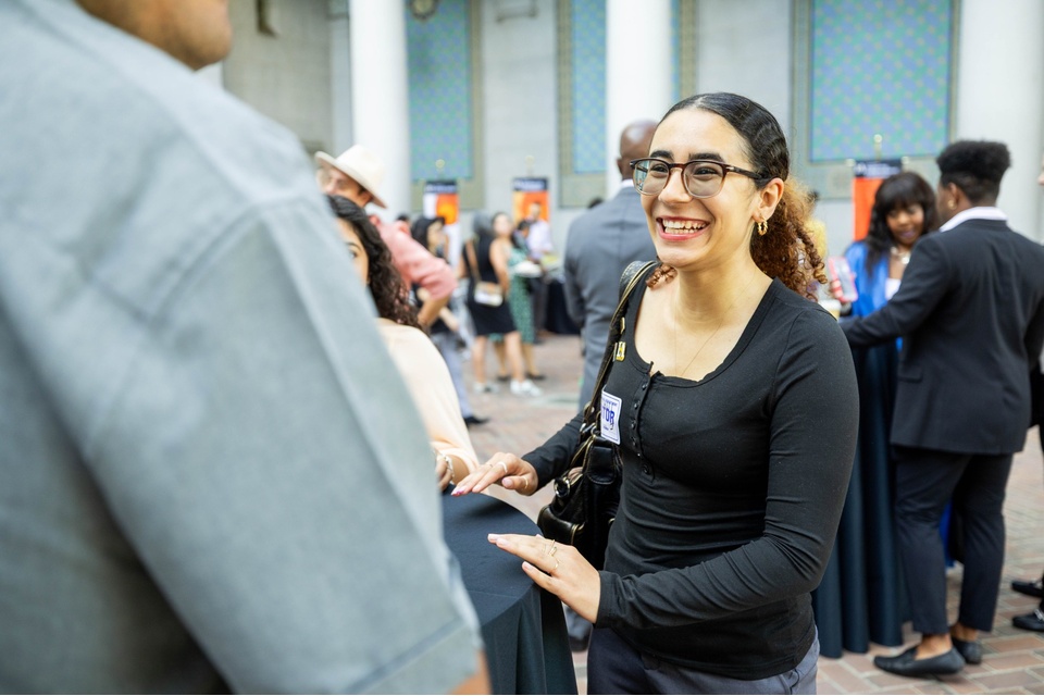 Cal State LA student smiles while networking at a professional event, speaking with other attendees in a large indoor space with columns and a patterned wall.