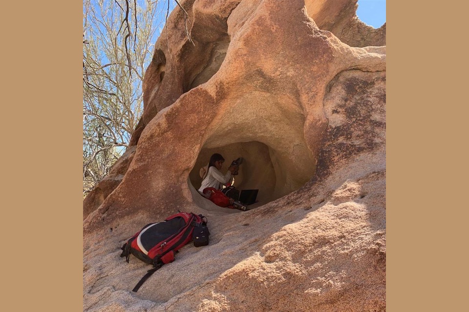 A field researcher works inside a small cave-like rock formation, with equipment and a backpack, surrounded by sunlit boulders and desert vegetation.