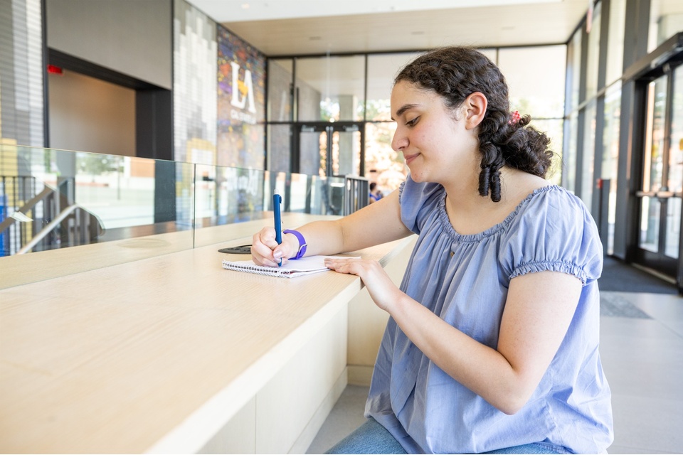 A Cal State LA student sits at a wooden counter inside a modern campus building, writing in a notebook with LA signage in the background.