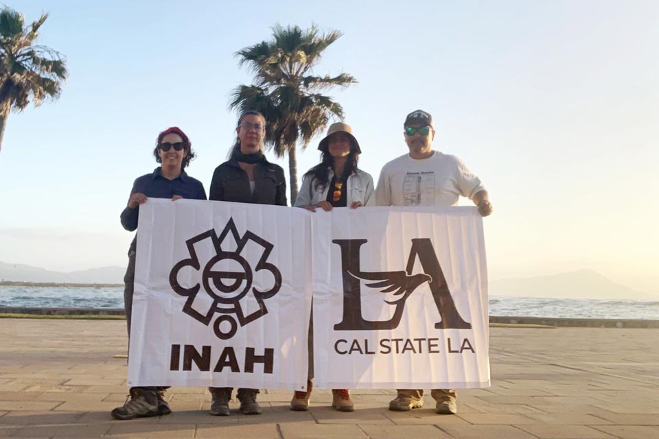Four people stand on a promenade by the ocean at sunset, holding banners for INAH and Cal State LA, with palm trees and mountains in the background.