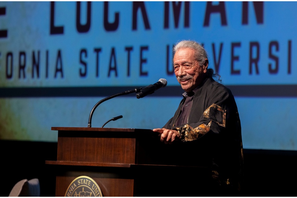 Edward James Olmos speaks at a podium on stage during an event at Cal State LA, smiling while addressing the audience.