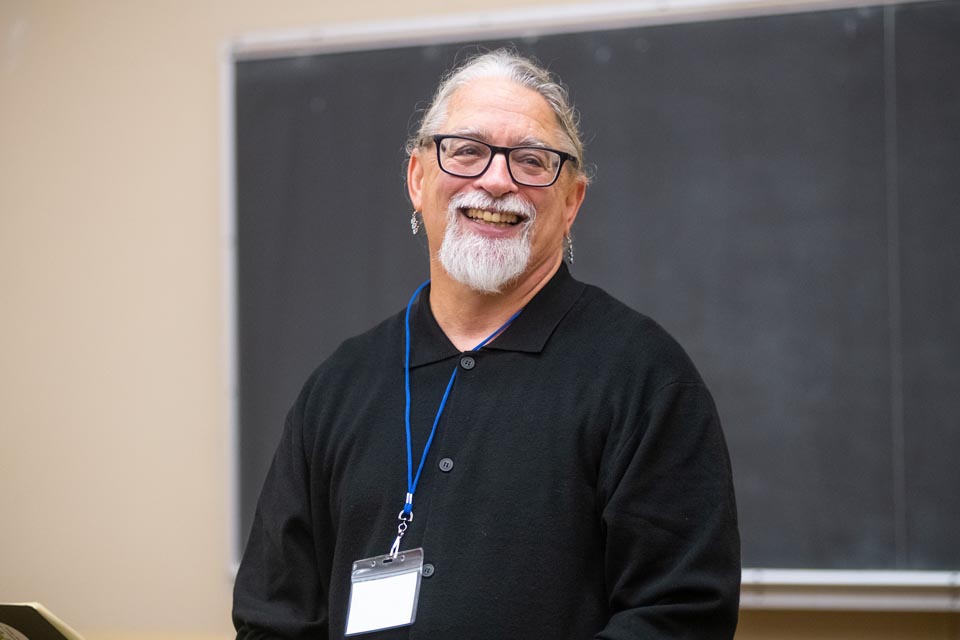 A faculty member with gray hair sits on a couch in an academic building, smiling warmly toward the camera.