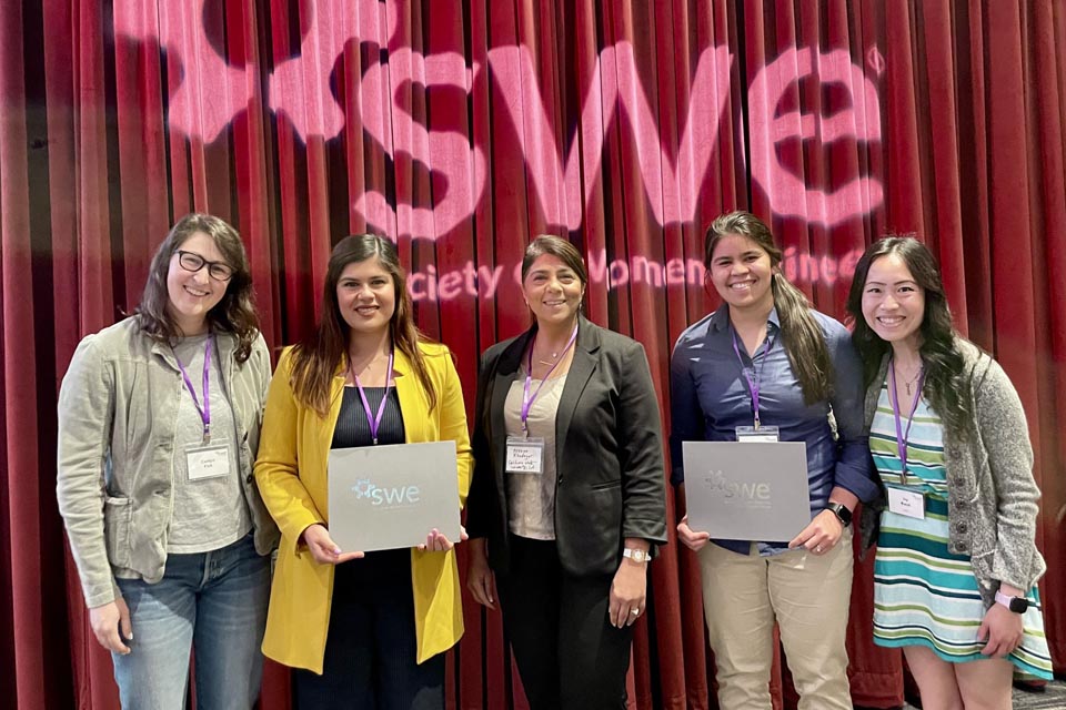 Five Society of Women Engineers members stand together smiling with SWE certificates in front of a red curtain and event backdrop.