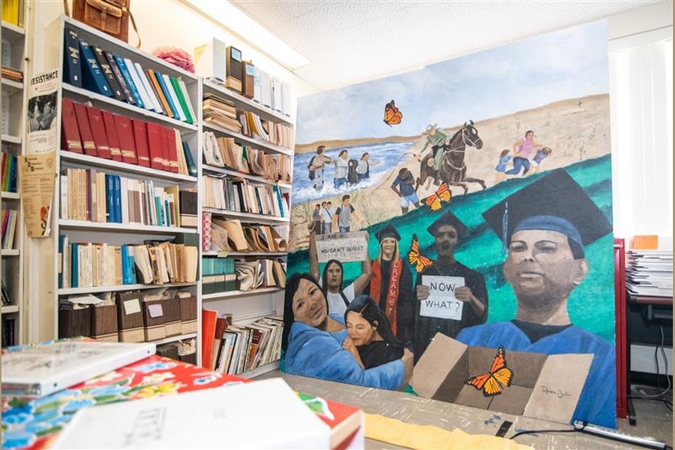 A large painted mural depicting migrant journeys, family unity, graduation, and monarch butterflies is displayed inside a crowded office with bookshelves filled with academic materials.