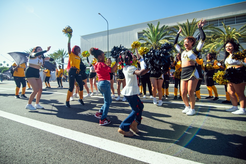 Cheerleaders and community members dance in the street during the MLK Day Parade, with palm trees and campus buildings in the background.