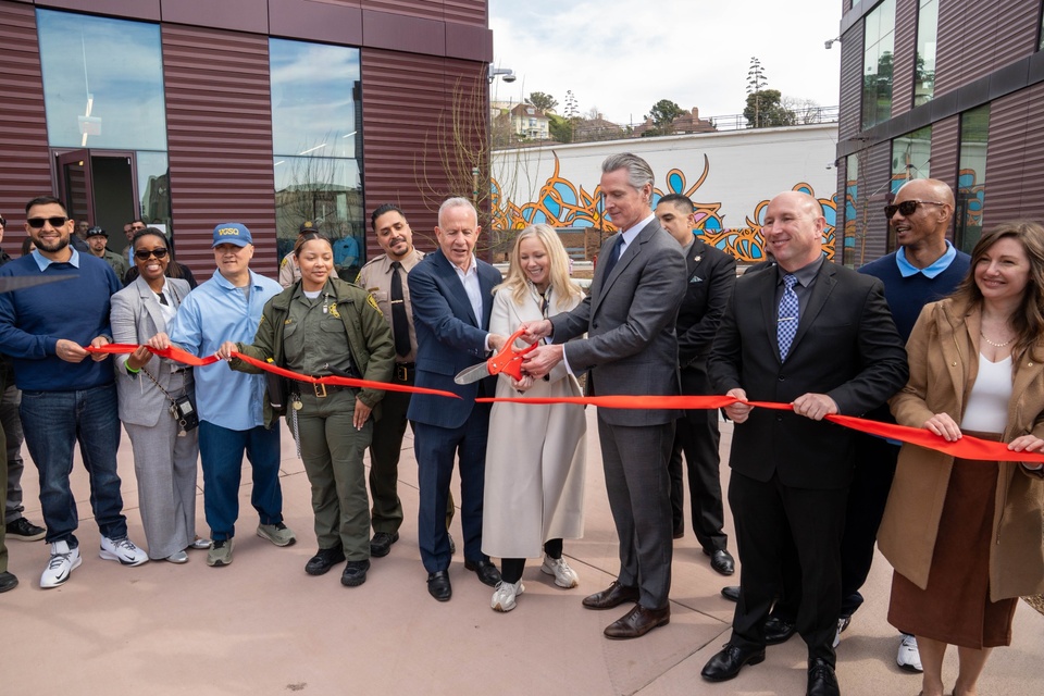 Group of officials and community members participate in a ribbon-cutting ceremony outside a modern building, with a red ribbon stretched across the front.