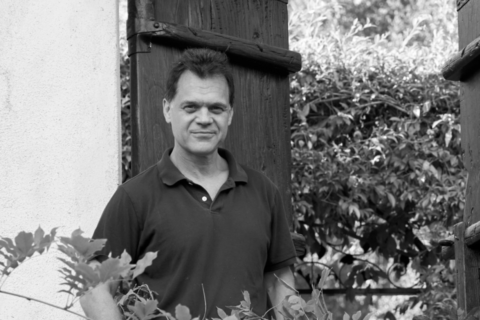 Black-and-white portrait of a man standing beside a wooden gate and stucco wall, surrounded by foliage.