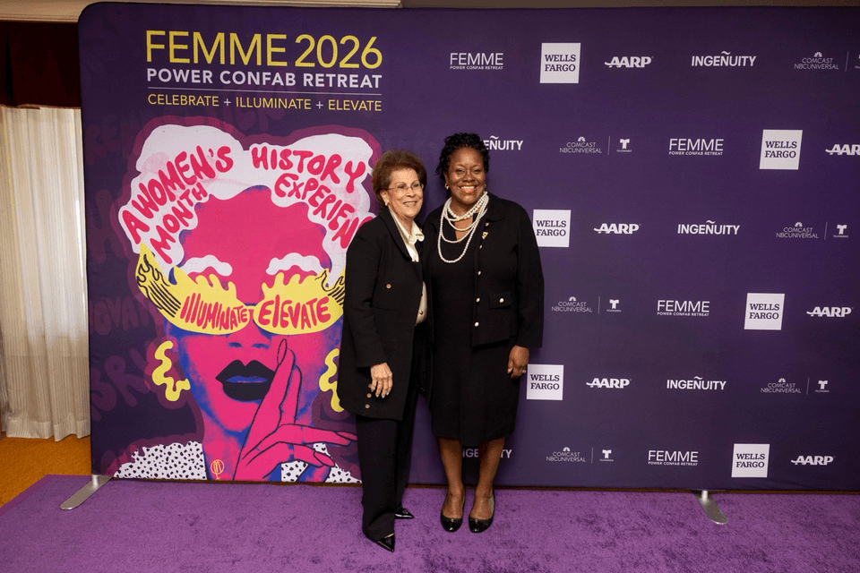 Two leaders pose together at the FEMME 2026 Power Confab Retreat in front of a purple backdrop with “Women’s History Experience” artwork.