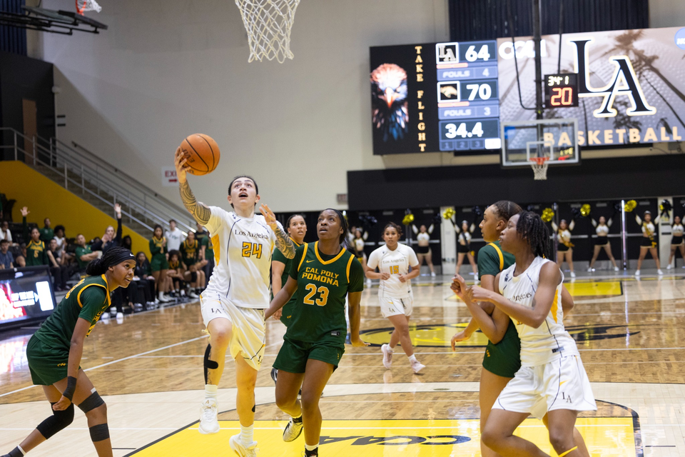 Cal State LA women’s basketball player drives to the basket during a game against Cal Poly Pomona as teammates and defenders watch under the scoreboard.