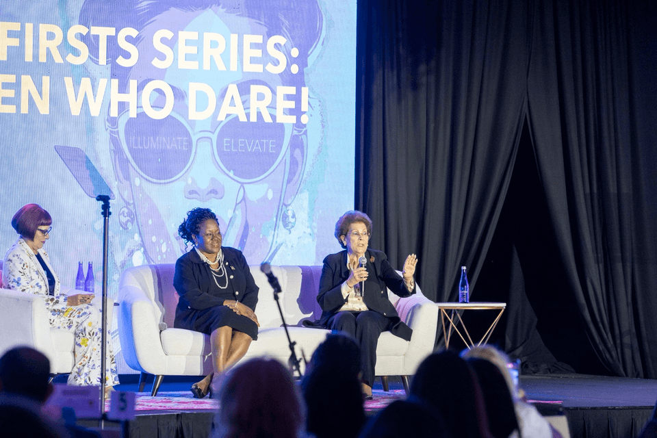 Panelists speak on stage during a FEMME event as audience members watch, with a large screen reading “Firsts Series: Women Who Dare!”