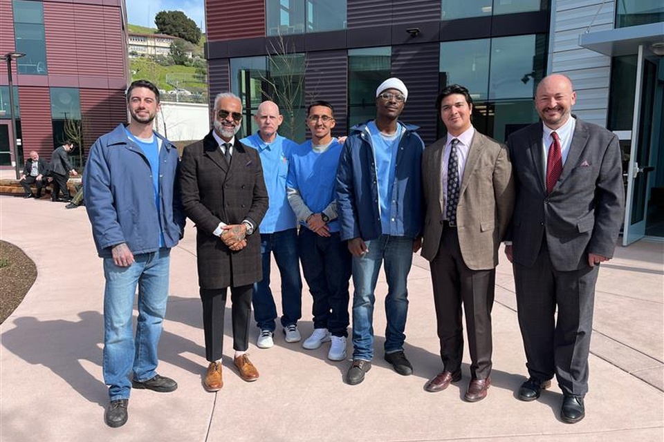Group of seven men standing outside a modern building at a learning center, posing together in daylight.