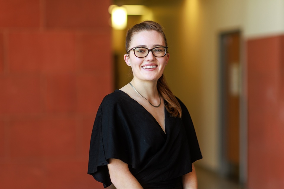Researcher smiles while standing in a campus hallway, wearing a black top and glasses at Cal State LA.