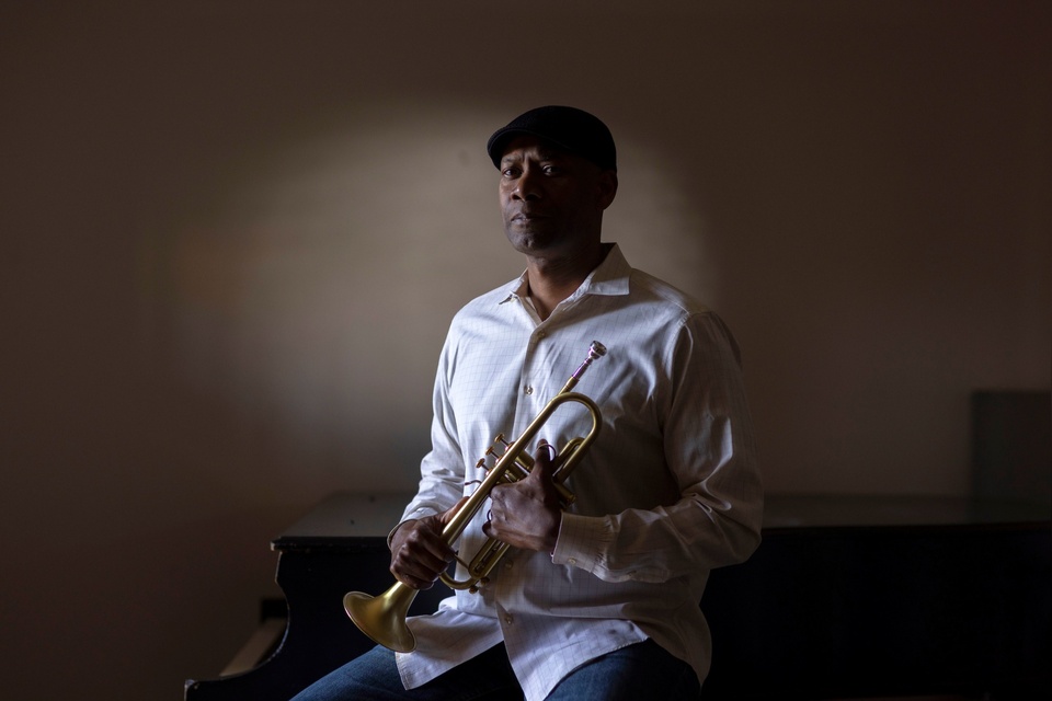 Musician holds a trumpet while seated beside a piano in a softly lit room.