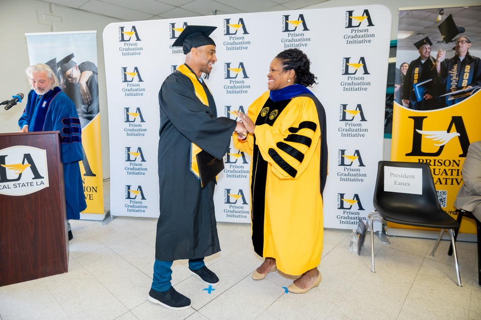 Cal State LA President Berenecea Johnson Eanes congratulates a graduate during a Prison Graduation Initiative ceremony as they shake hands in front of a program backdrop. 