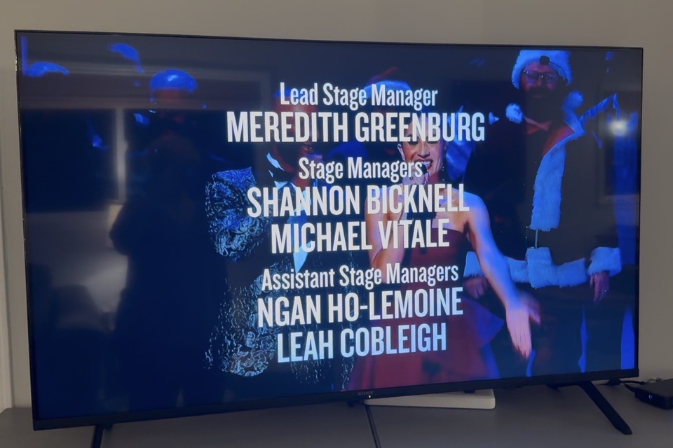 Television screen displays stage management credits over an image from a theatrical production.
