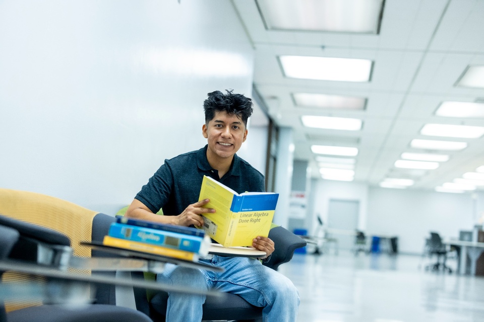 Student sits in a campus study area holding a linear algebra textbook, with additional math books in the foreground.