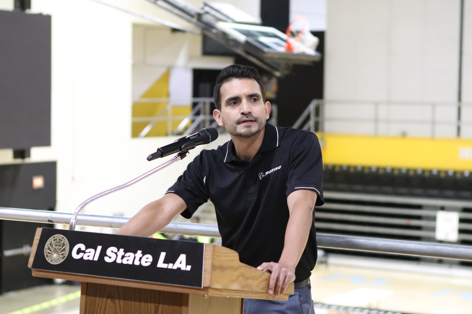 A speaker wearing a Boeing polo addresses an audience from a Cal State LA lectern in a gymnasium.